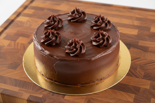 bright fun photo of 6 in round layer cake with chocolate frosting on top of cake are 5 swirls of chocolate frosting edge of cake icing is smooth cake on top of gold cake board on top of a nice wooden kitchen cutting board straight on view