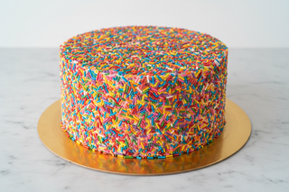 bright photo of a round cake completely covered in rainbow sprinkles, straight on view and the cake is on a gold cake board and on a marble counter